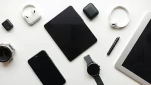 Flat lay of various modern tech gadgets including smartphone, smartwatch, wireless earbuds, and tablet arranged on a minimalist desk with neutral background