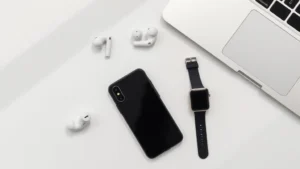 Flat lay of modern tech gadgets including smartphone, wireless earbuds, smartwatch, and laptop on white minimalist surface with soft shadows
