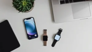 Top-down flat lay of modern tech gadgets: smartphone, wireless earbuds, smartwatch, and laptop arranged on minimalist desk with neutral background, natural lighting emphasizing materials and design