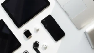 Flat lay of various modern tech gadgets arranged on a white surface: smartphone, smartwatch, wireless earbuds, tablet, laptop corner visible, natural soft lighting from above