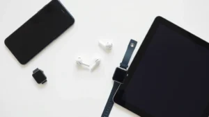 Overhead flat lay of various tech gadgets including smartphone, wireless earbuds, smartwatch, and tablet arranged neatly on a clean white surface with natural lighting